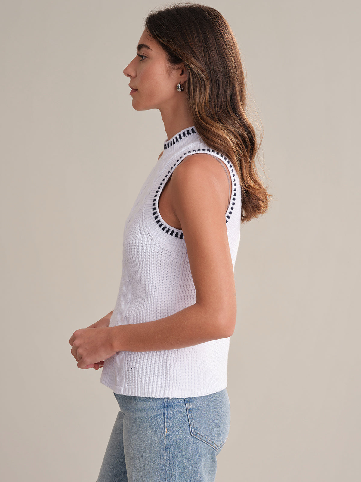 A woman stands in profile against a plain background, wearing a 525 America sleeveless white sweater tank with black double cable stitch trim and light blue jeans. Her hands are lightly clasped in front of her.
