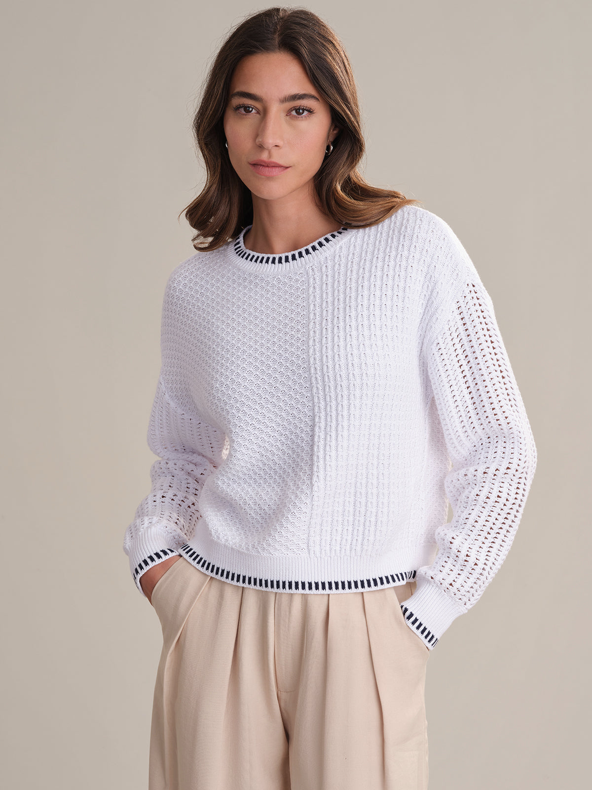 A woman models a 525 America lightweight cable knit sweater with contrast tipping on the crewneck and black-trimmed cuffs, paired with beige wide-leg pants. She stands with hands in pockets against a simple background.