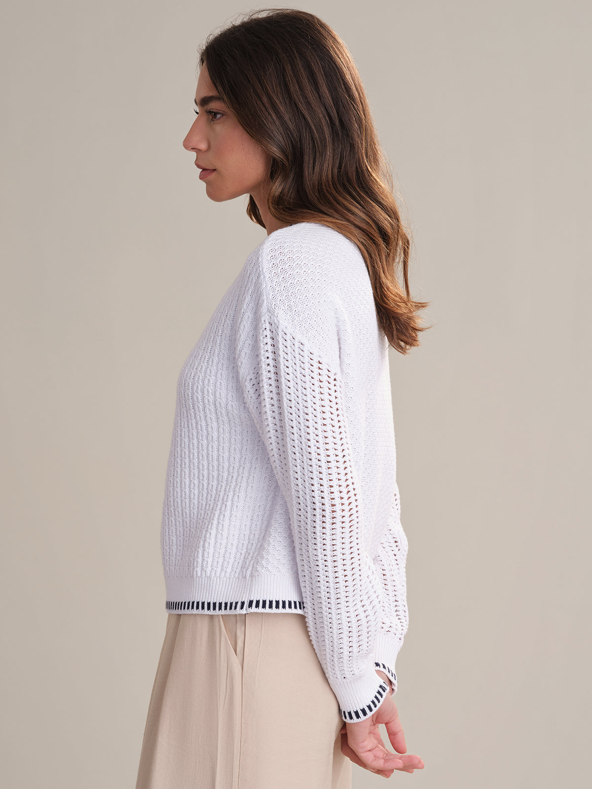 A woman with long brown hair stands in profile against a plain background, wearing a 525 America relaxed fit crewneck sweater with contrast tipping and patterned sleeves, paired with light beige pants. Her arms rest at her sides.