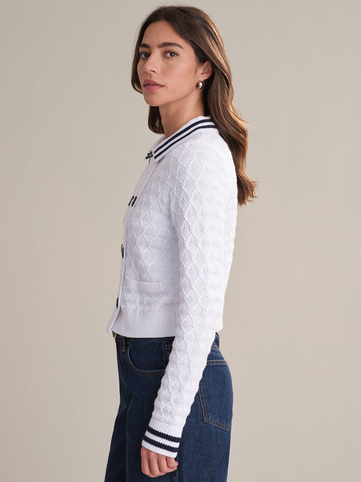 A woman with long brown hair stands in profile against a plain background, wearing a 525 America sailor-inspired white textured cardigan with navy trim and dark blue jeans.