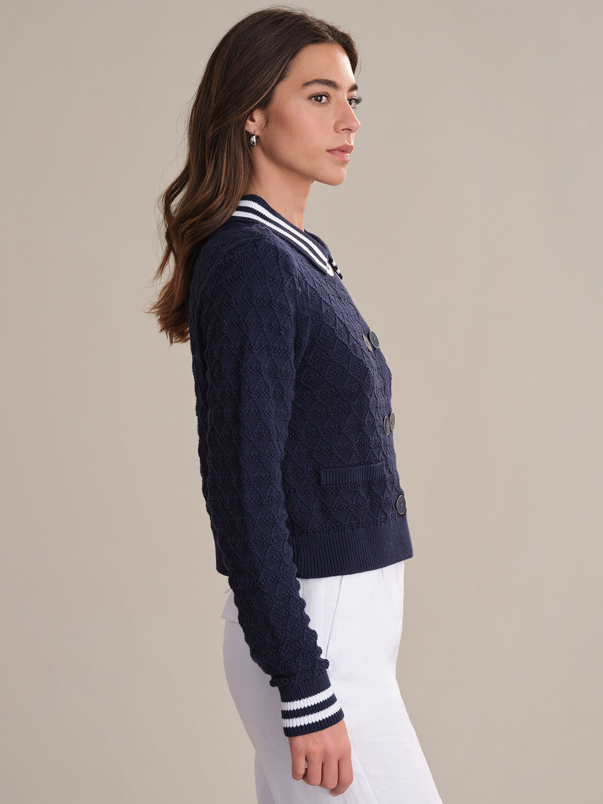 A woman with long brown hair stands in profile wearing a 525 America navy blue textured sailor-inspired cardigan with white striped cuffs and collar, paired with white pants, against a plain beige background.