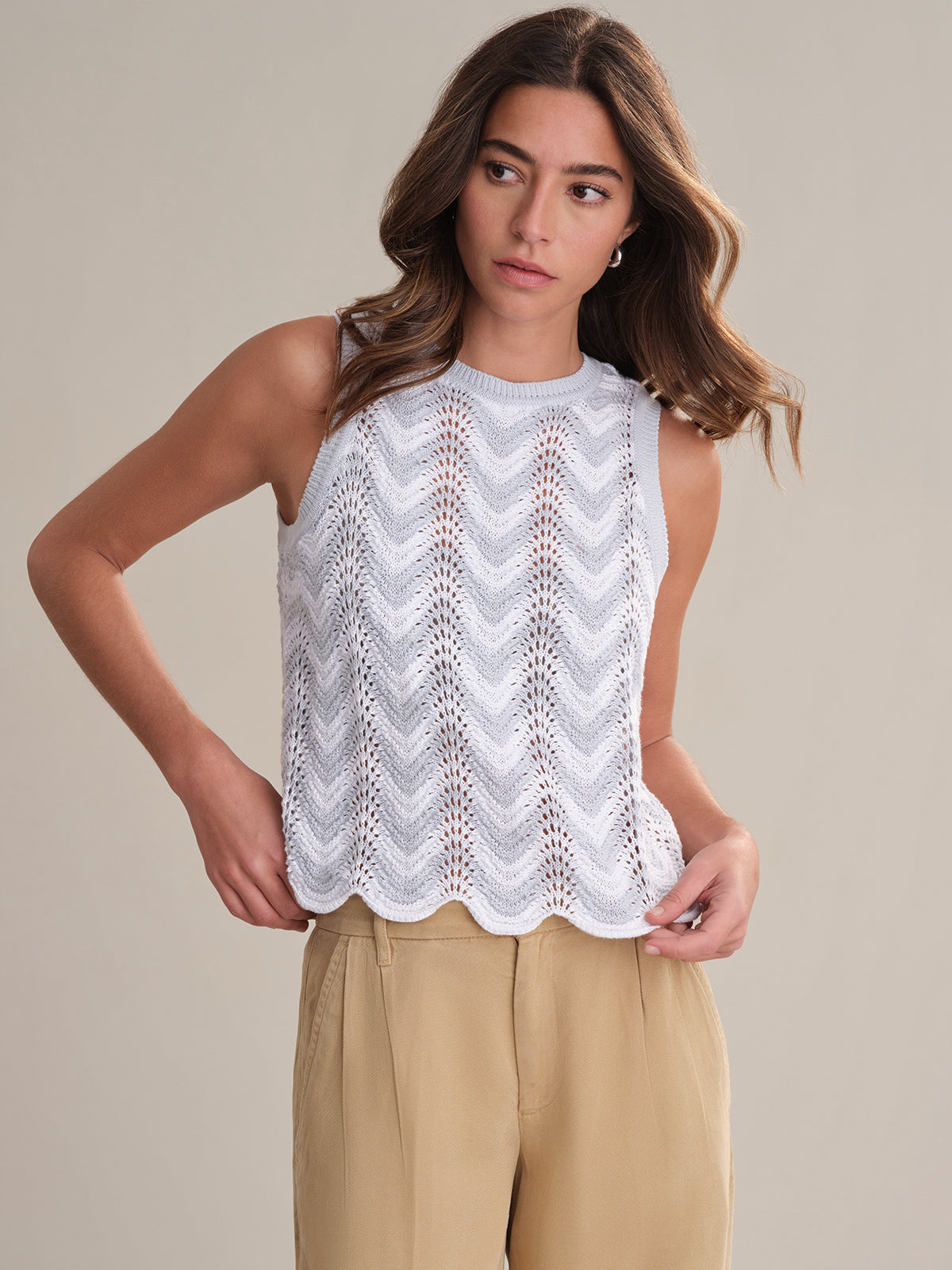 A woman with long brown hair stands against a plain background wearing a sleeveless white chevron pointelle knit top by 525 America and beige pants, gazing slightly to the side.
