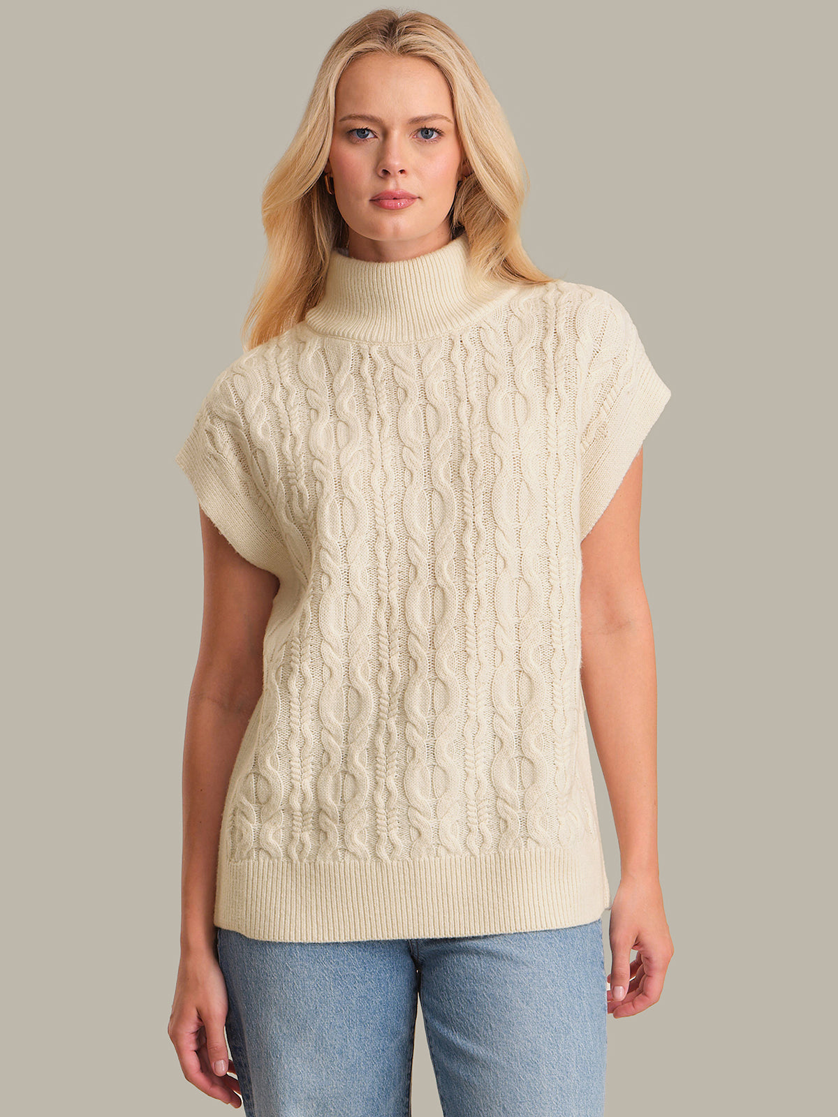 Woman wearing a cream cable knit sleeveless sweater against a neutral background