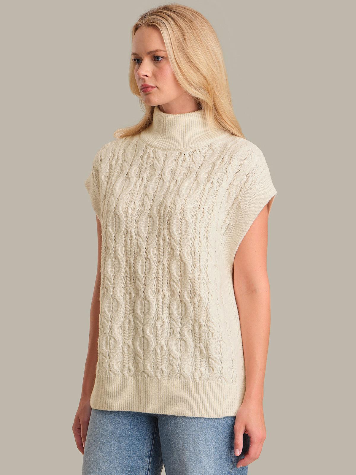 Woman wearing a beige cable knit sleeveless sweater against a plain background