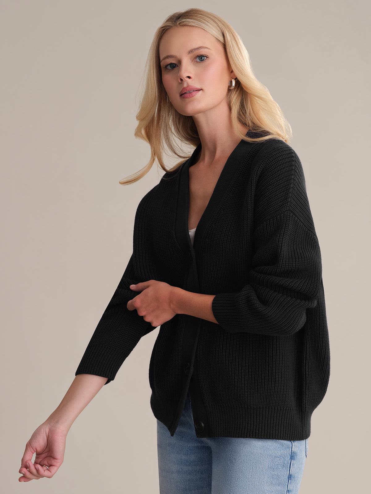 A person with shoulder-length blonde hair wears the oversized Emilia: Cotton Cocoon Cardigan in black over a white top and blue jeans. Against a neutral backdrop, they glance to the side with a calm expression, effortlessly exemplifying the art of layering.