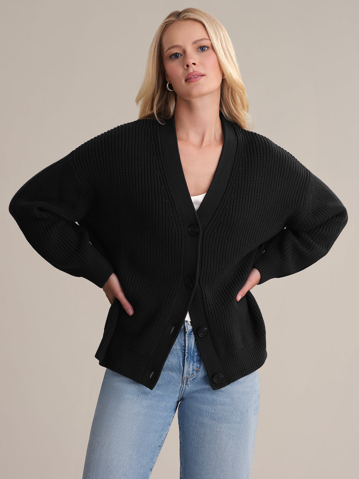 A person stands confidently with hands on hips, showcasing the Emilia: Cotton Cocoon Cardigan in an oversized fit. Made from 100% cotton, its layered over a white top and light blue jeans against a plain beige background, highlighting a casual, stylish look perfect for effortless layering.