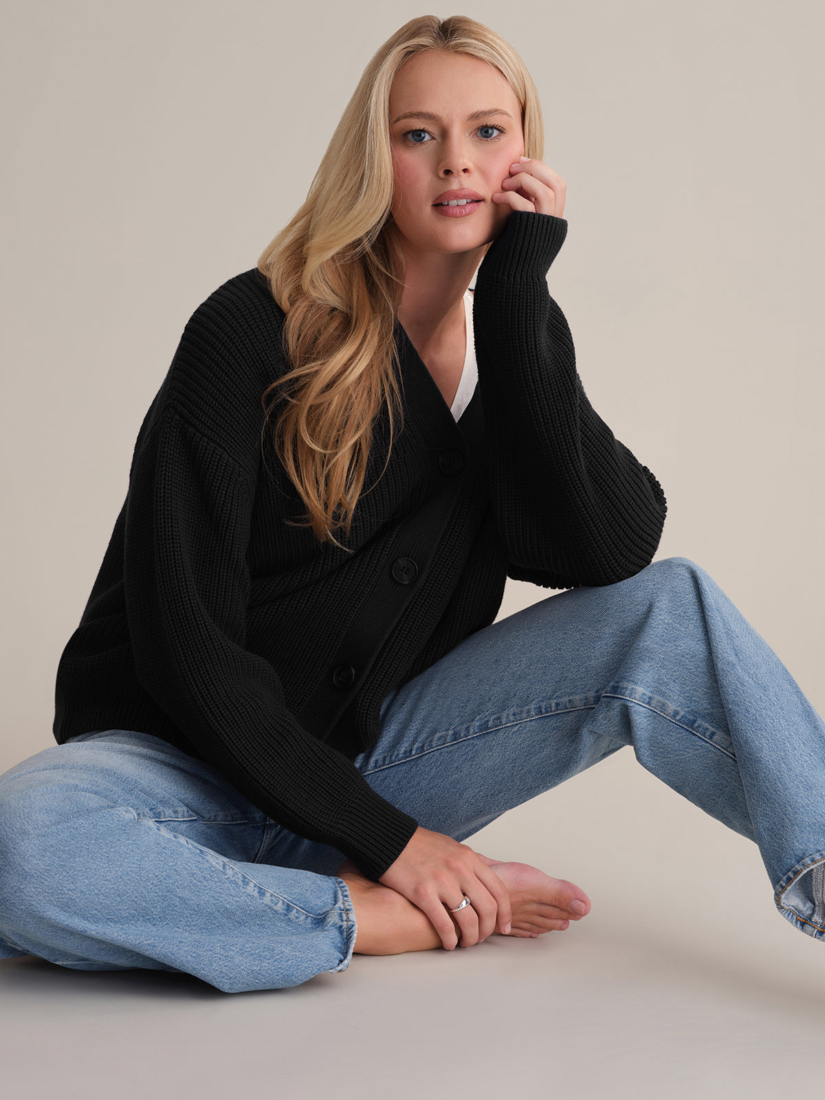A person with long hair sits on the floor against a neutral backdrop, wearing an oversized Emilia: Cotton Cocoon Cardigan in black and blue jeans. Their cheek rests on their hand as they gaze forward with a relaxed expression, one knee bent and the other leg extended.