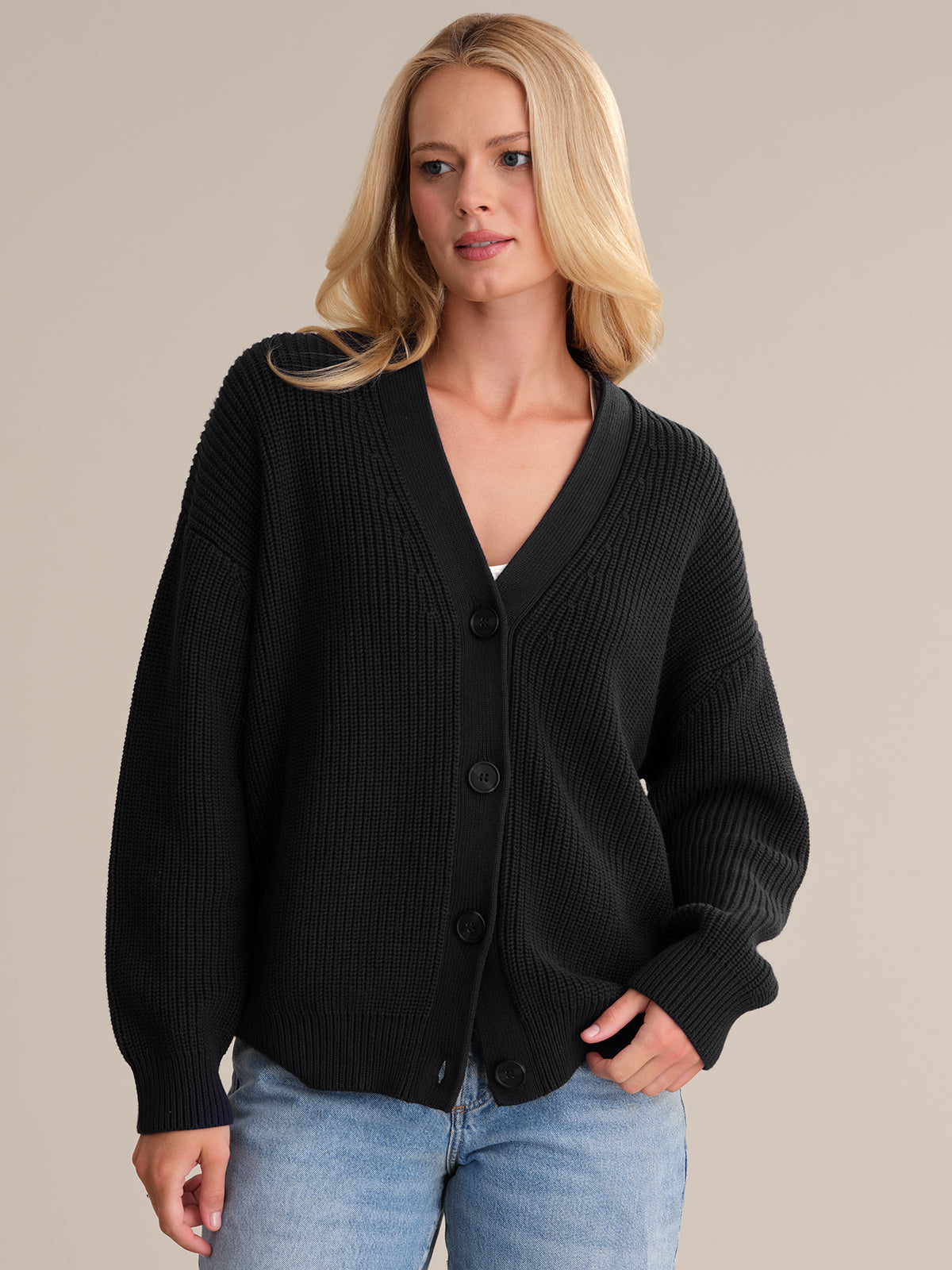 Wearing the Emilia: Cotton Cocoon Cardigan in oversized black, paired with light blue jeans, a person stands against a neutral background. Made from 100% cotton, the look radiates comfort and style. With long blonde hair and a relaxed pose, they casually rest one hand in their pocket.