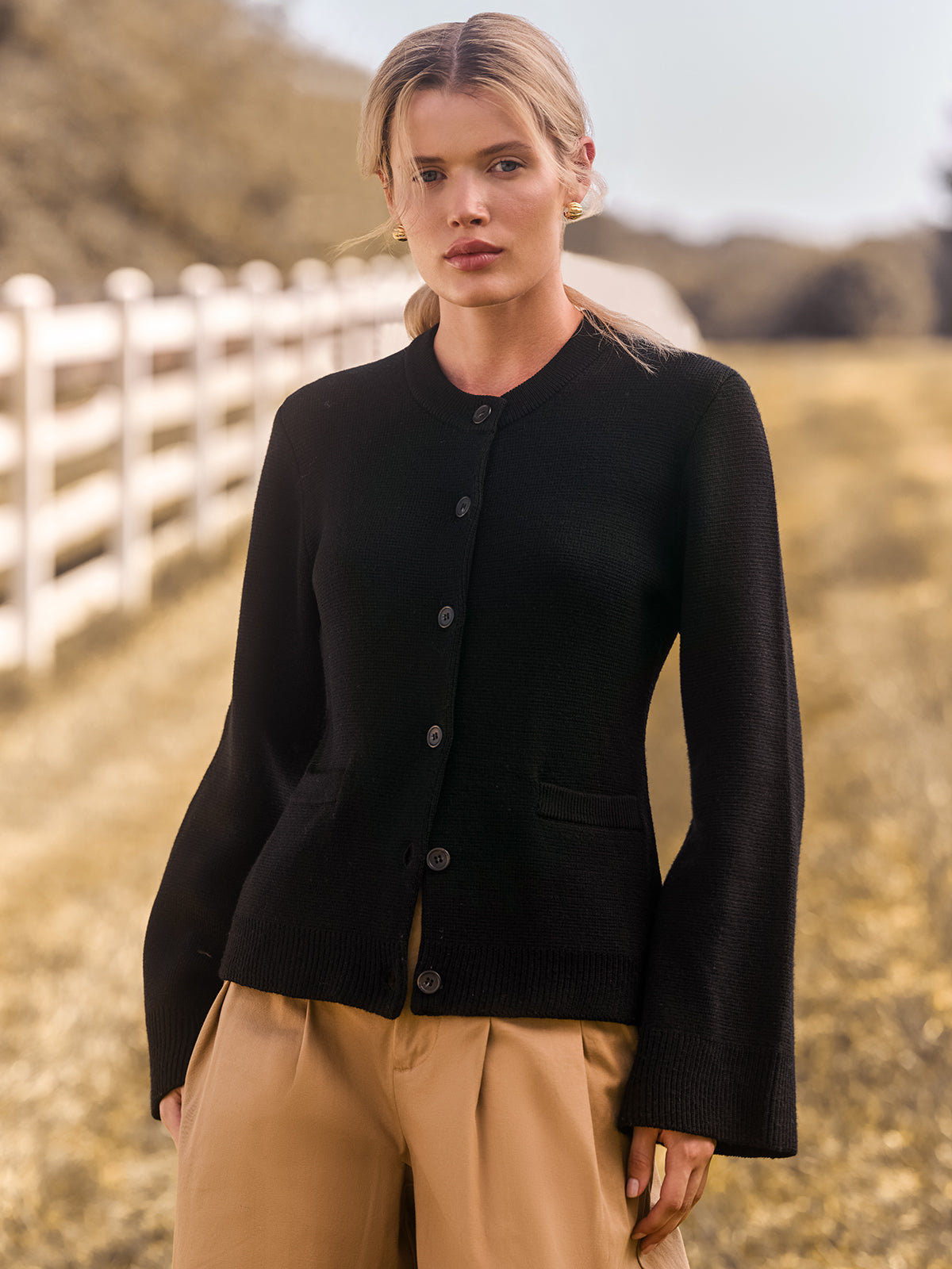 Woman wearing a black cardigan and beige pants standing in a field with a white fence in the background.