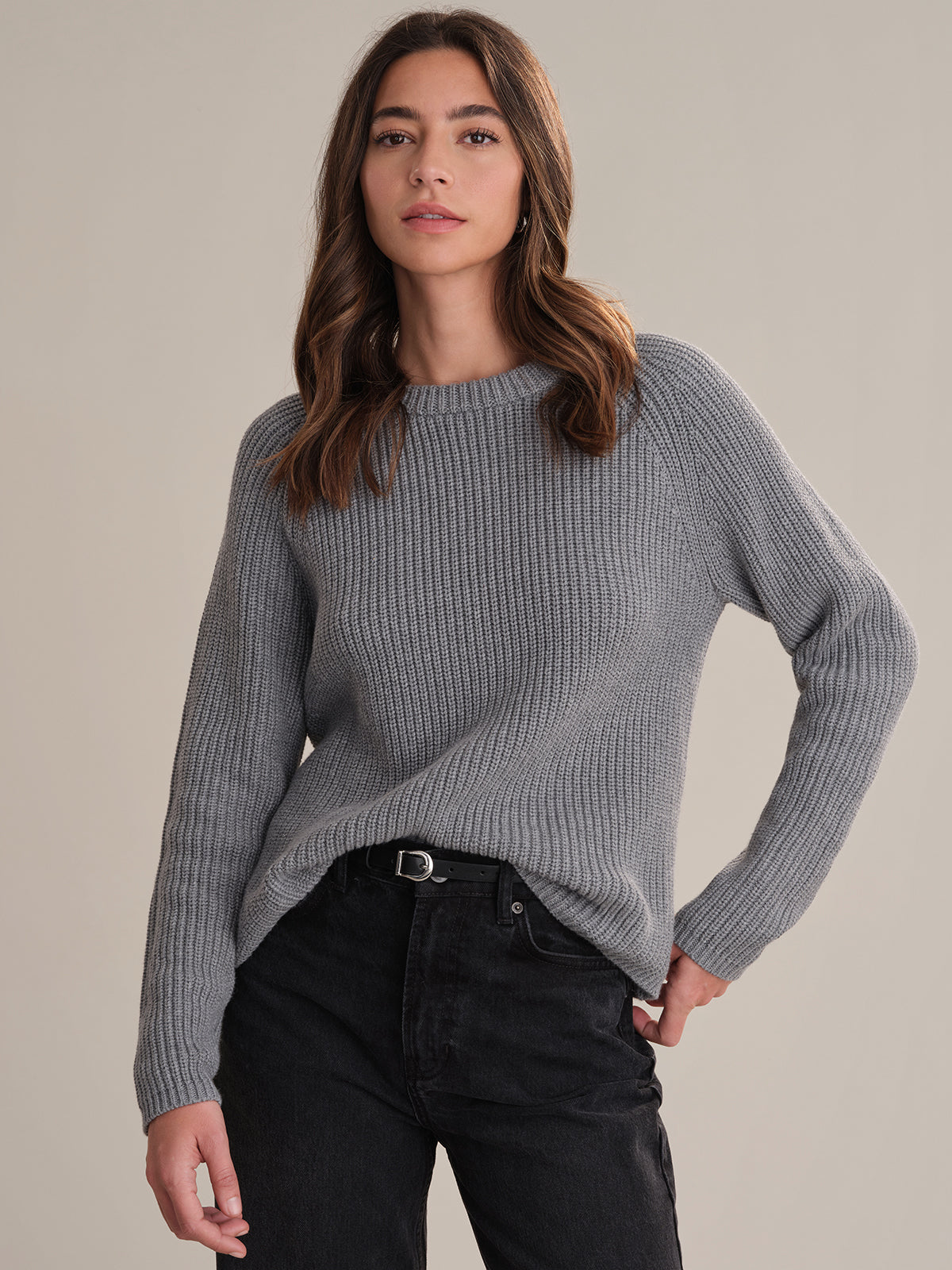 A person with long brown hair wears a 525 America gray raglan sleeve sweater and black jeans, standing against a beige background with one hand on their hip.