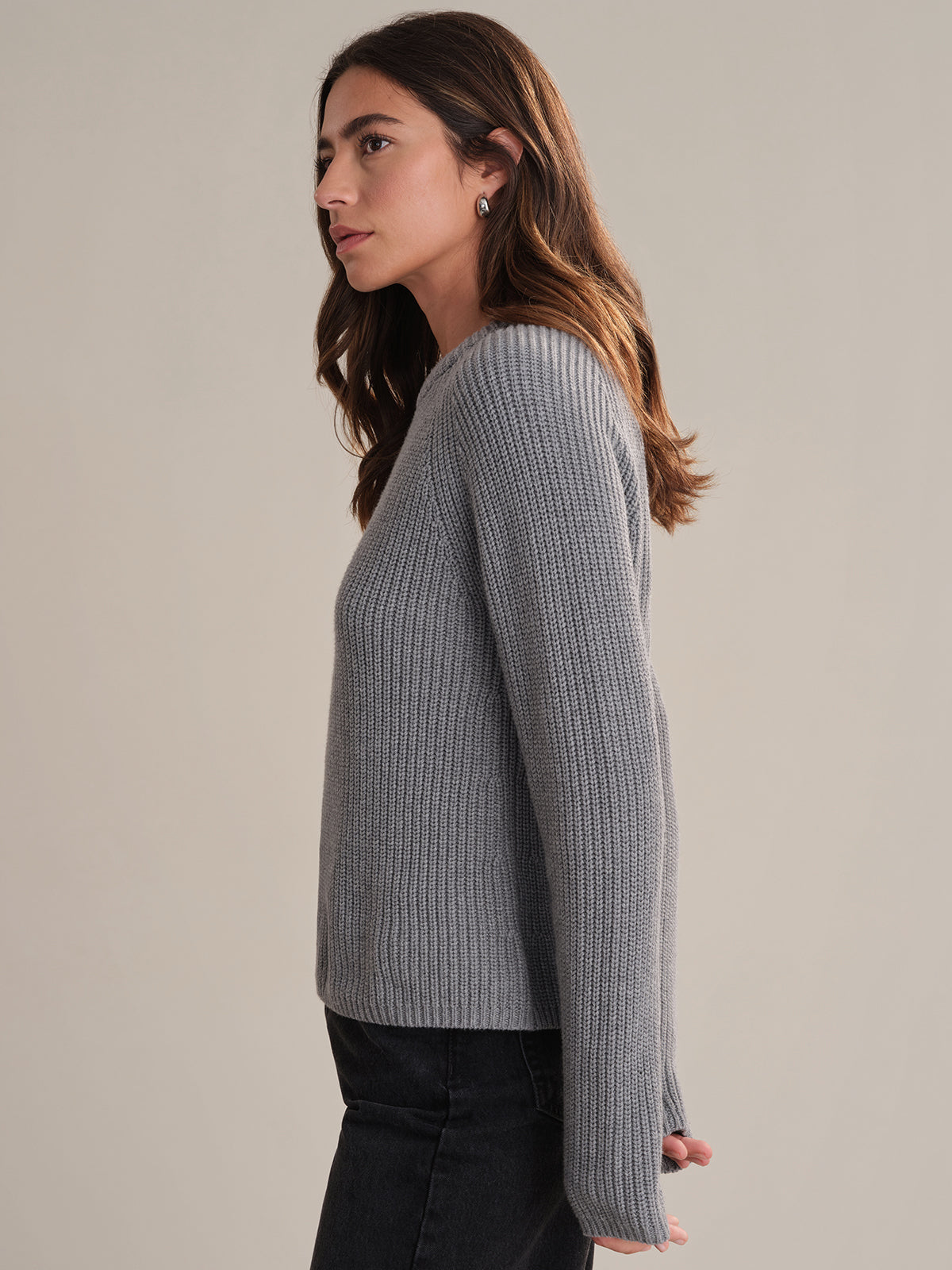 A woman with long brown hair stands in profile against a plain background, wearing a gray 525 America cotton shaker stitch sweater with a fitted silhouette and black pants. Her arms are relaxed at her sides as she looks slightly ahead.