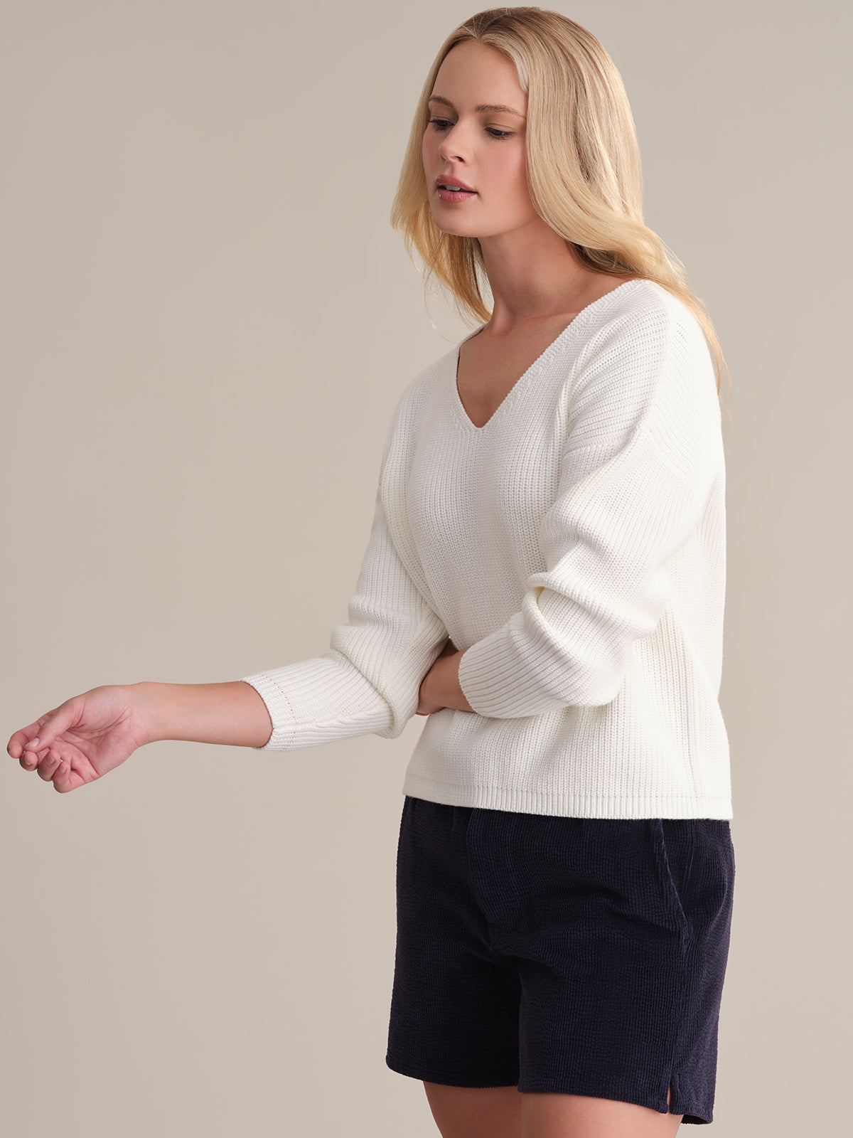 A person with long blonde hair wears a 525 America Ava V-Neck Sweater in a shaker stitch design. The 100% cotton sweater is paired with dark shorts, contrasting against the plain beige background as they stand with one arm slightly extended.