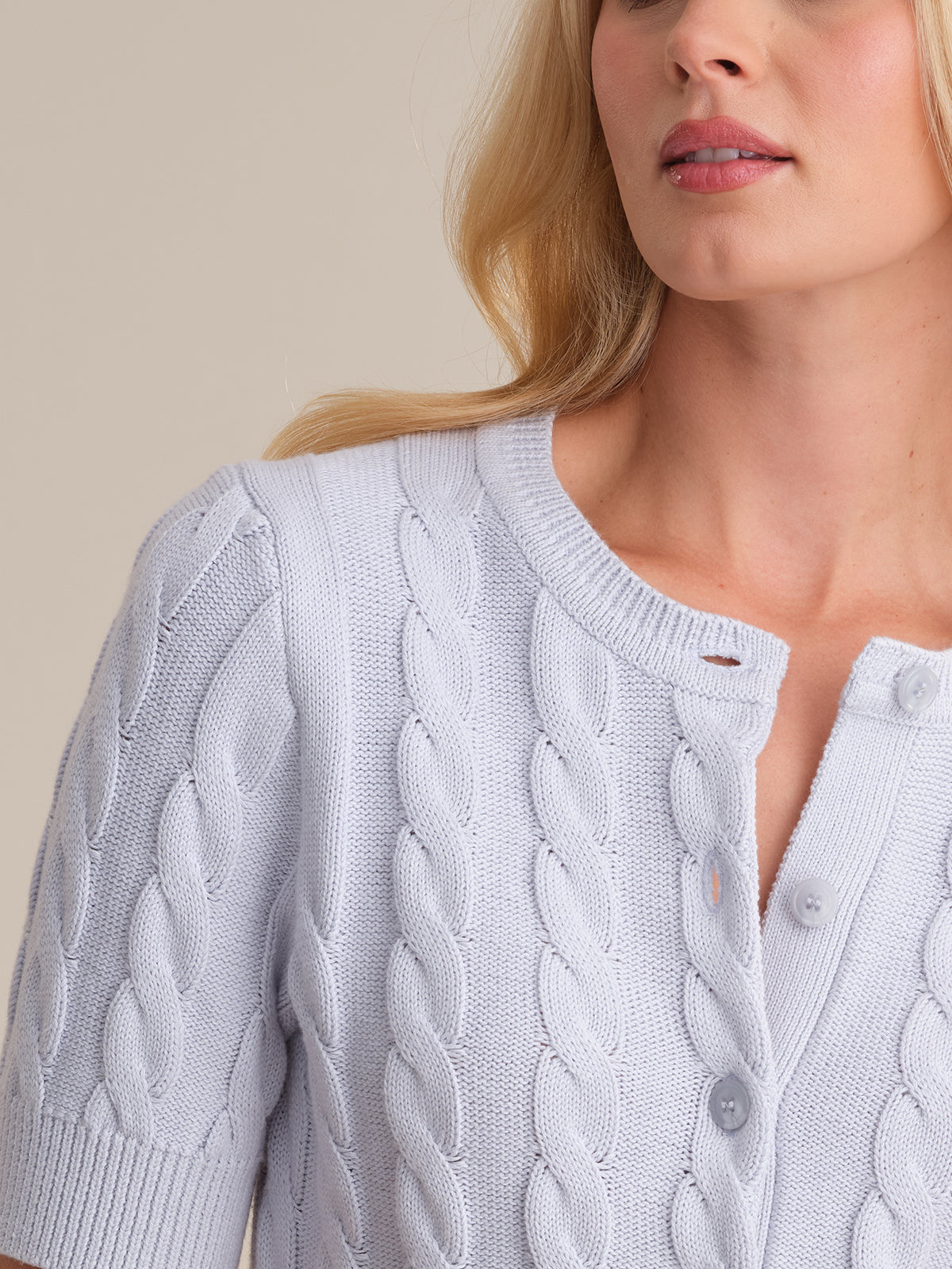 A close-up of a woman wearing a 525 America 100% cotton cable cardigan with short sleeves and large buttons. Her face is partially visible, and her long blonde hair drapes over her shoulder.