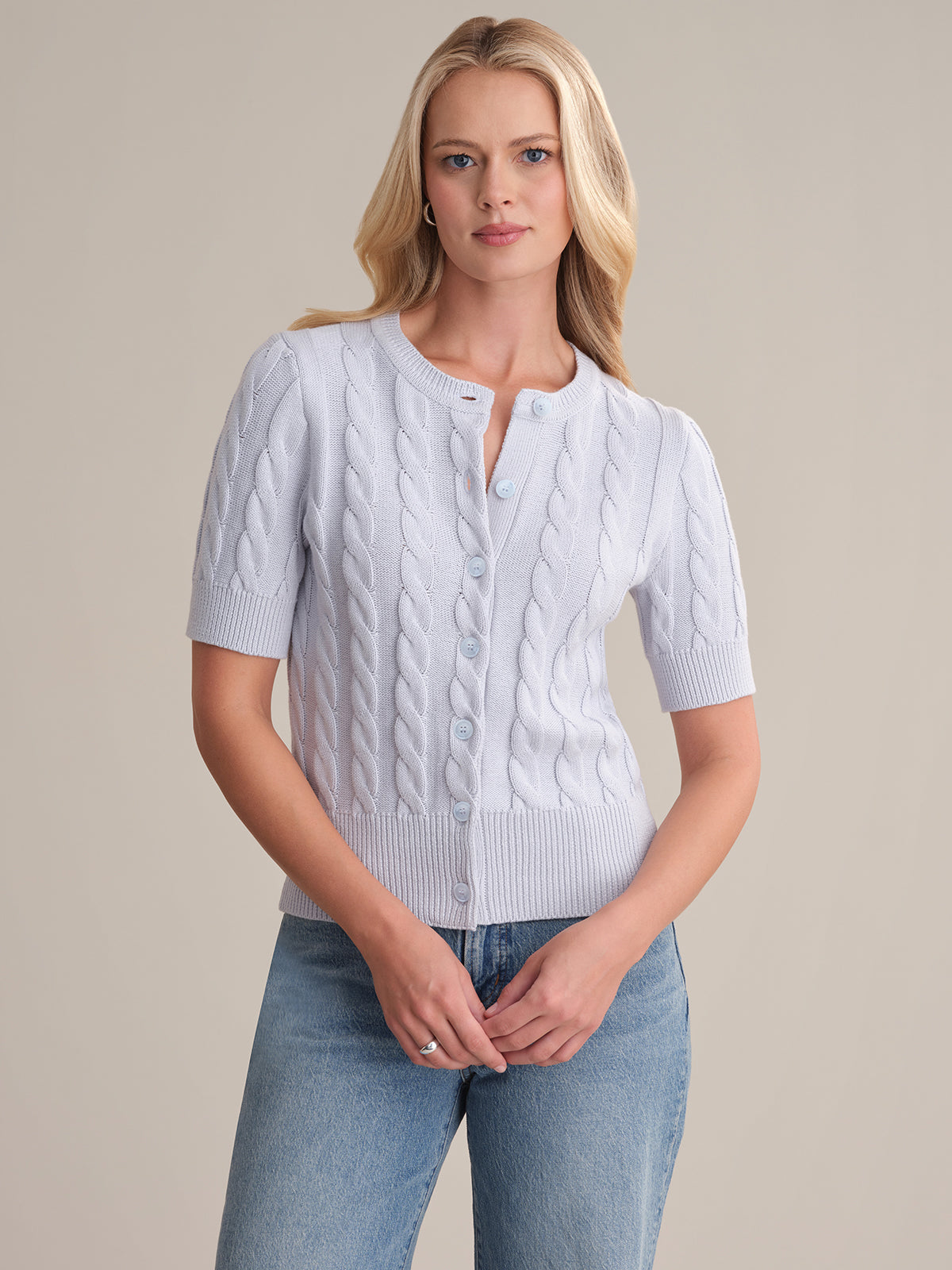 A woman with long blonde hair wears a 525 America 100% cotton short-sleeve cable cardigan with buttons and blue jeans, standing against a plain light background and looking straight at the camera.