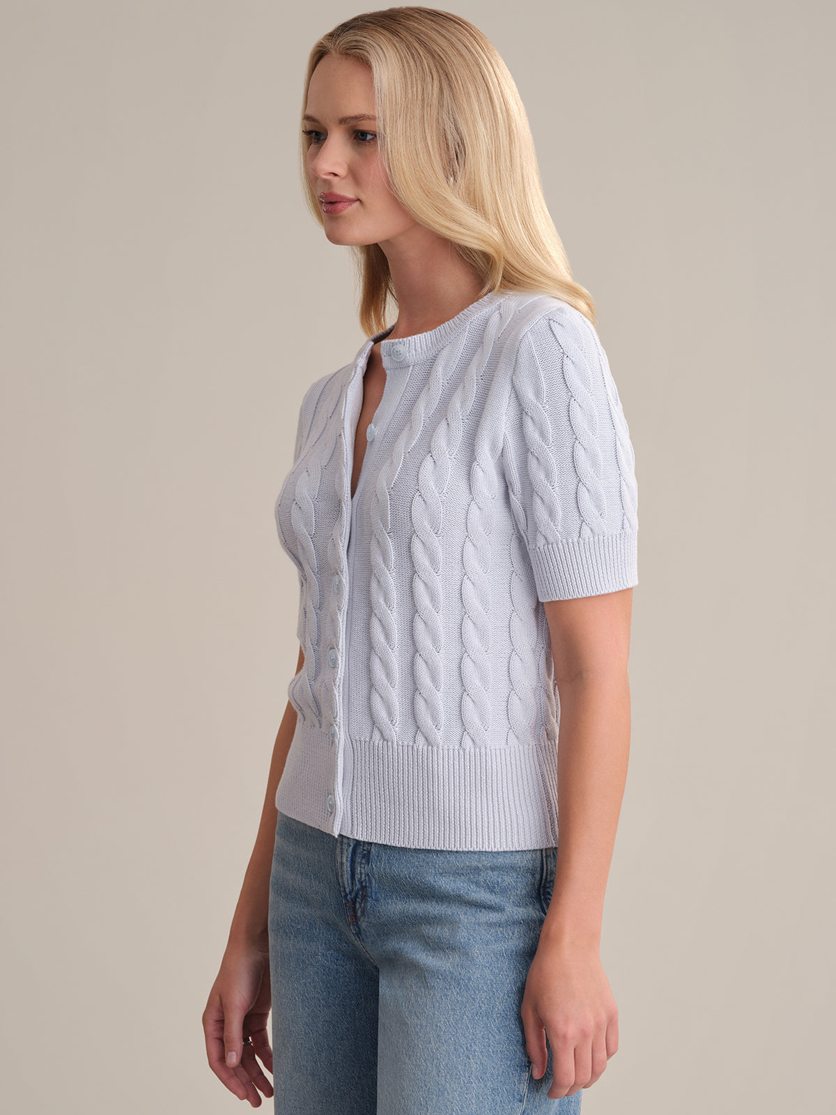 A woman with long blonde hair stands facing slightly right against a neutral background, wearing a light blue 100% cotton cable cardigan with short sleeves from 525 America over blue jeans.