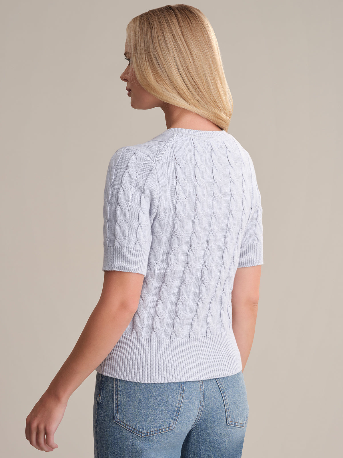 A person with light blonde hair is seen from behind wearing a 525 America light blue, 100% cotton cable knit short sleeve sweater and blue jeans, standing against a plain beige background.