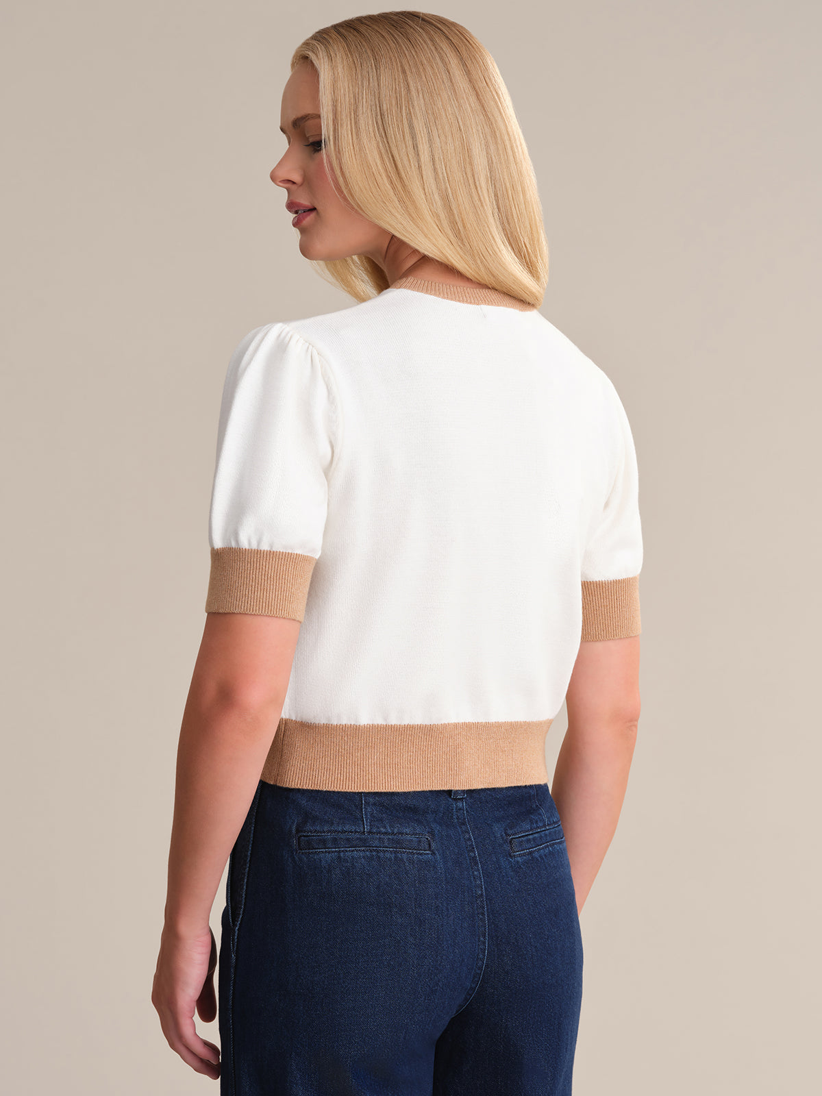 The individual with long blonde hair is wearing a 525 America white short-sleeve pullover made of 100% cotton, featuring tan ribbed cuffs and hem. Its paired stylishly with dark blue jeans against a neutral backdrop.