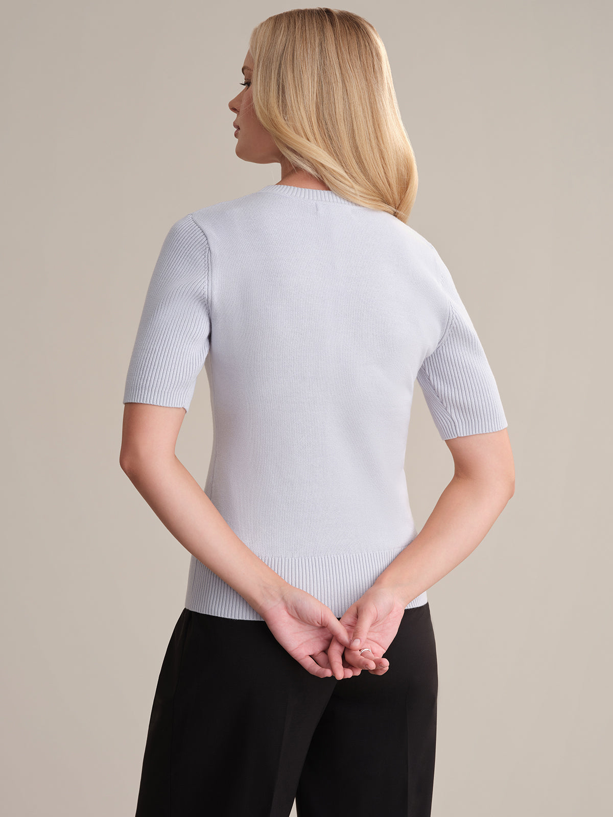 A person with long blonde hair, wearing a 525 America Short Sleeve Cotton Sweater (100% cotton) and black pants, stands facing away from the camera with hands clasped behind their back—an effortless everyday look.