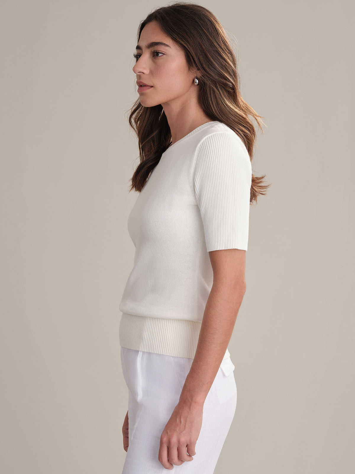 A woman with long brown hair stands in profile against a plain background, wearing a fitted 100% cotton short-sleeve top by 525 America and white pants—an effortless addition to your everyday wardrobe. She looks ahead with a neutral expression.