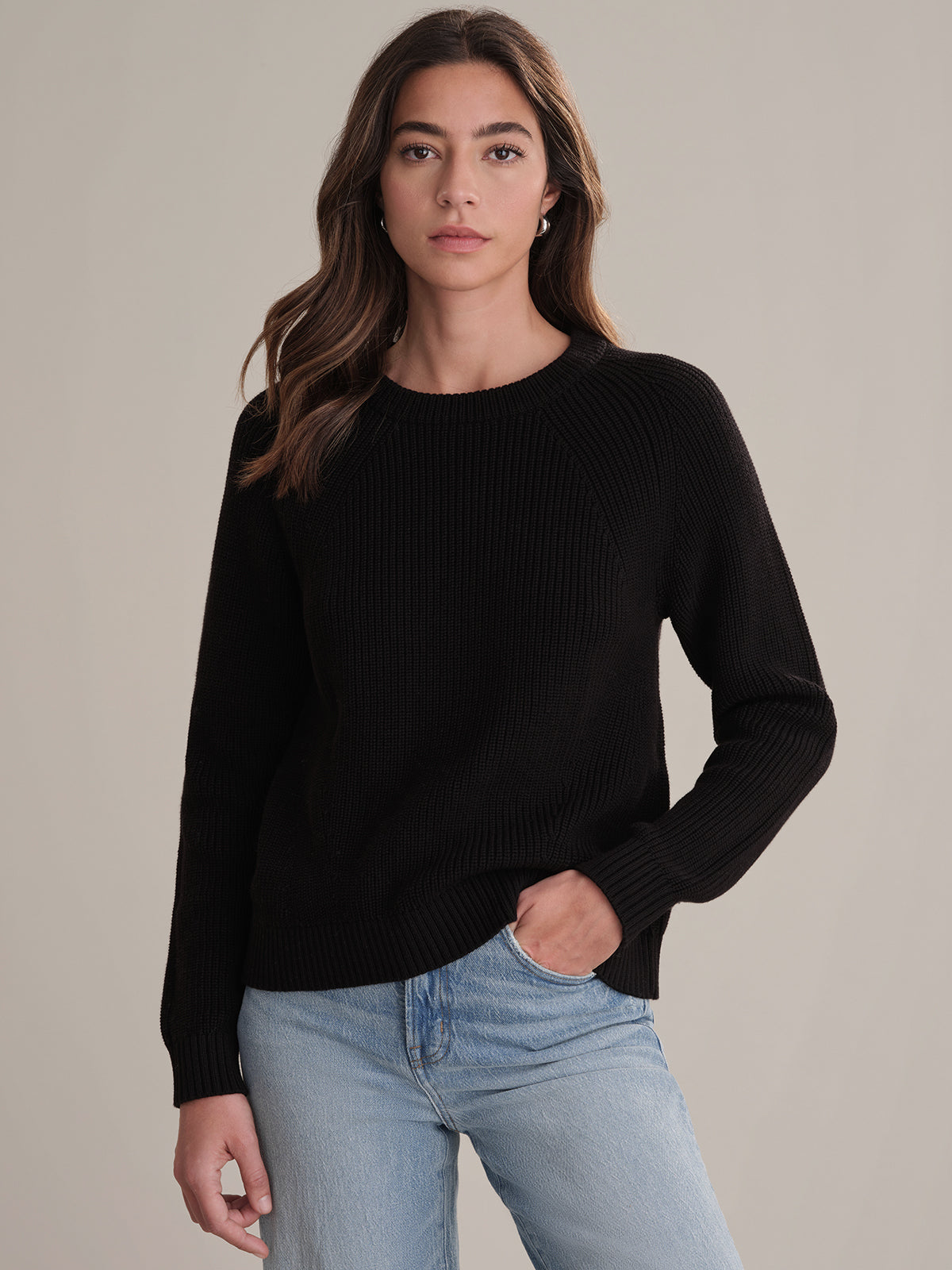 A person with long brown hair wears a black shaker stitch cotton sweater by 525 America and light blue jeans, standing against a plain beige background and looking at the camera with a neutral expression.
