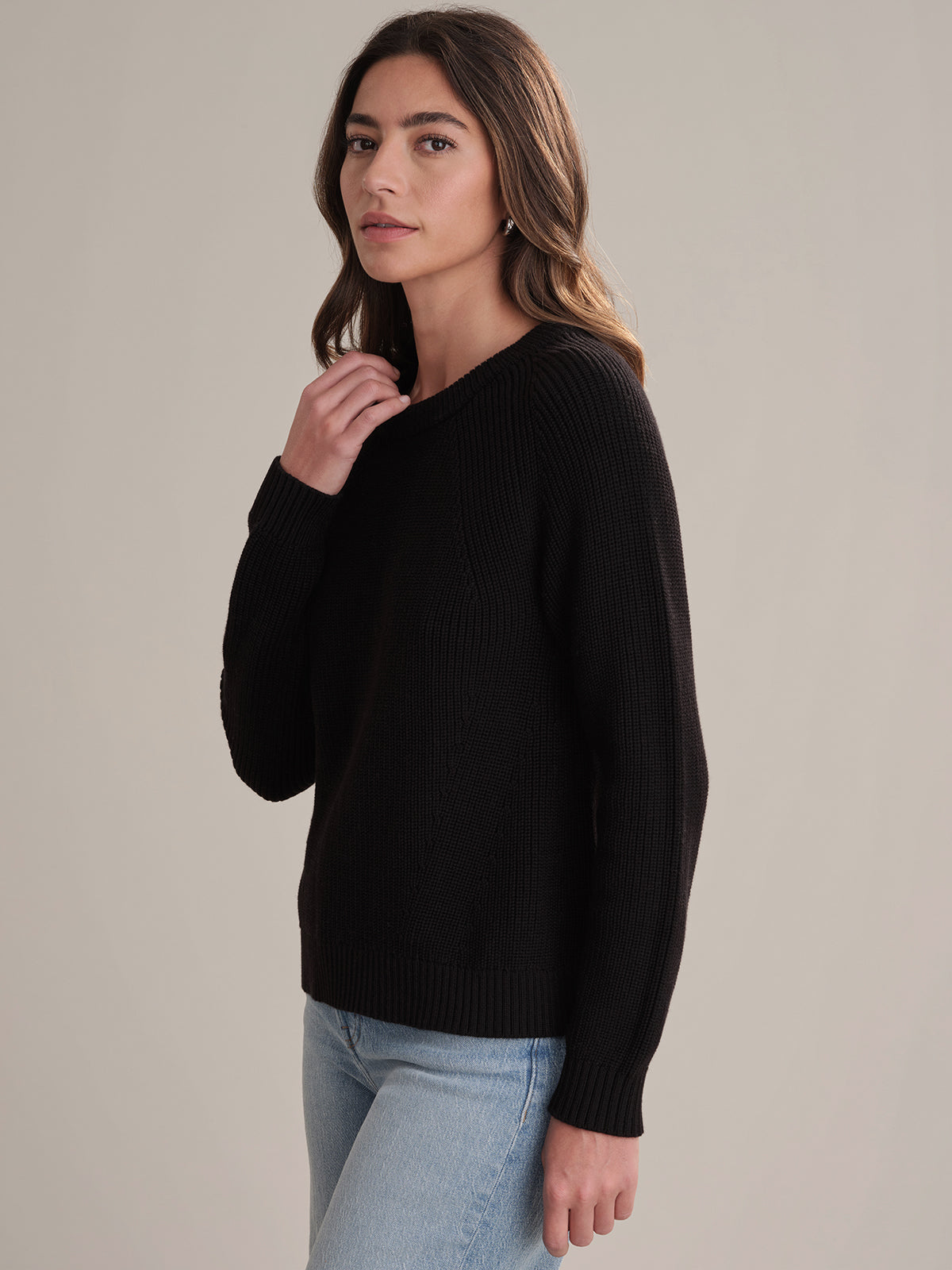 A woman with long brown hair wears a 525 America black shaker stitch sweater with raglan sleeves and light blue jeans, standing before a neutral background and looking at the camera with a neutral expression.