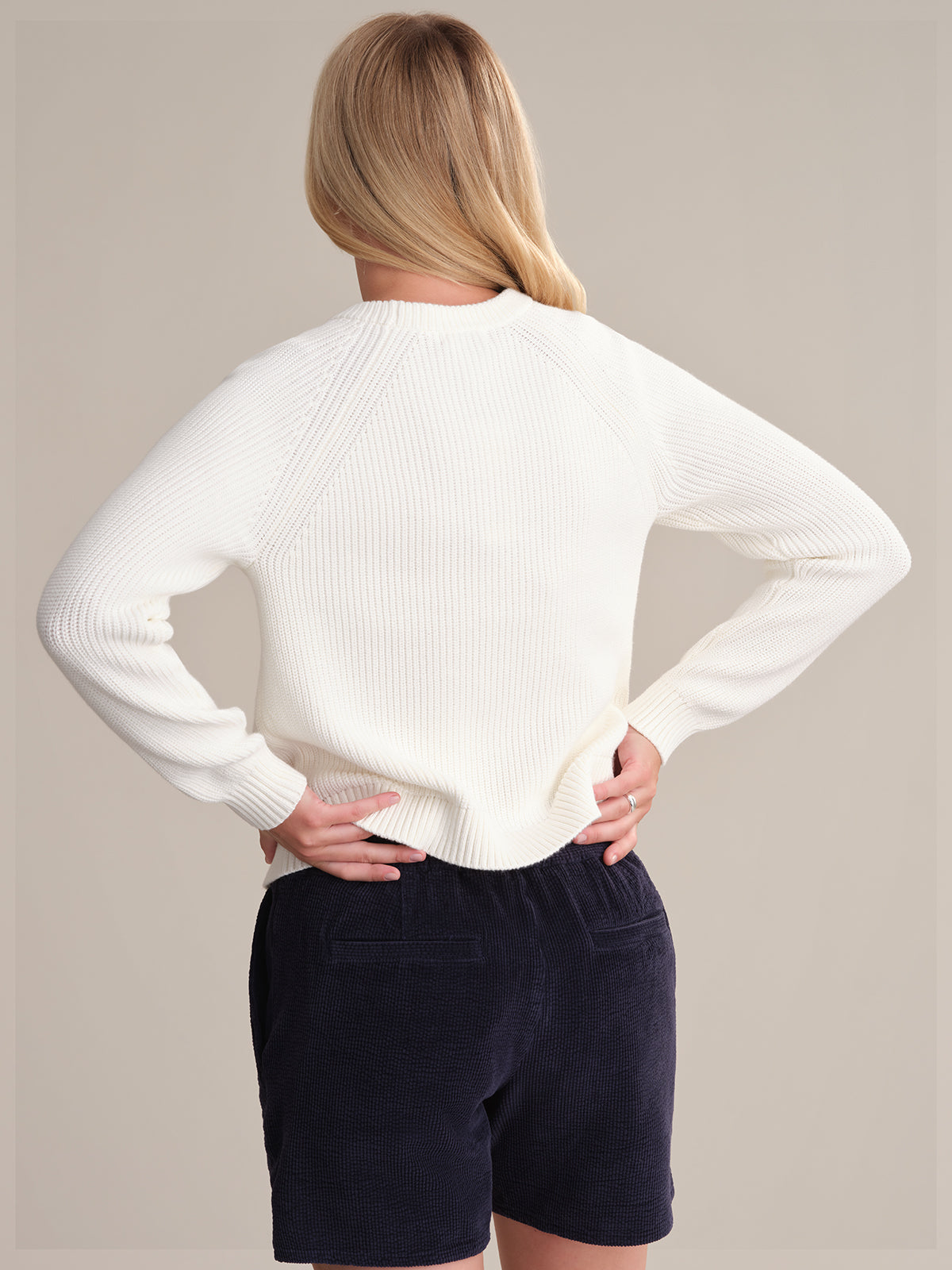A person with blonde hair, seen from the back, wears a fitted white 525 America cotton shaker stitch sweater and dark blue corduroy shorts. Their hands rest on their hips against a plain, light background.