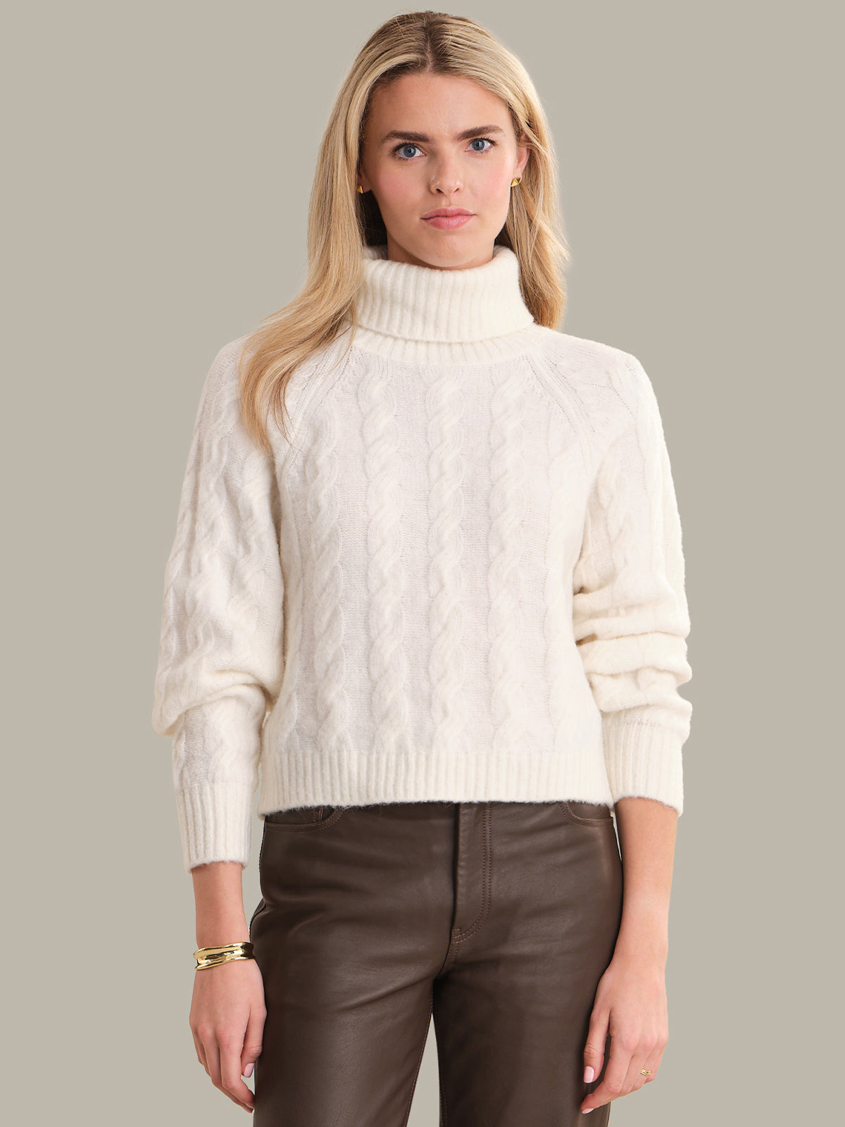 Woman wearing a white turtleneck sweater and brown pants against a gray background