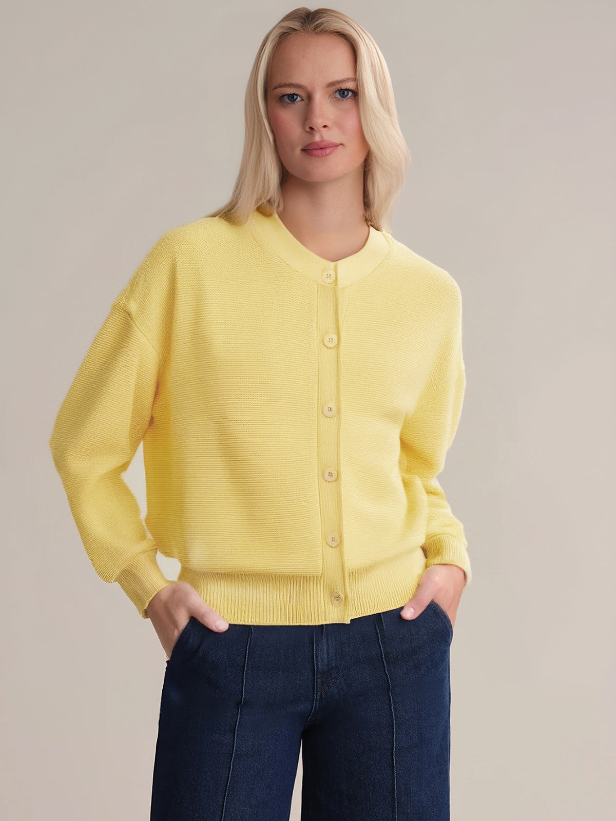 A person with long blonde hair wears a 525 America yellow lightweight knit button-up cardigan with a crew neckline and dark blue jeans, standing with hands in pockets against a plain, light background.