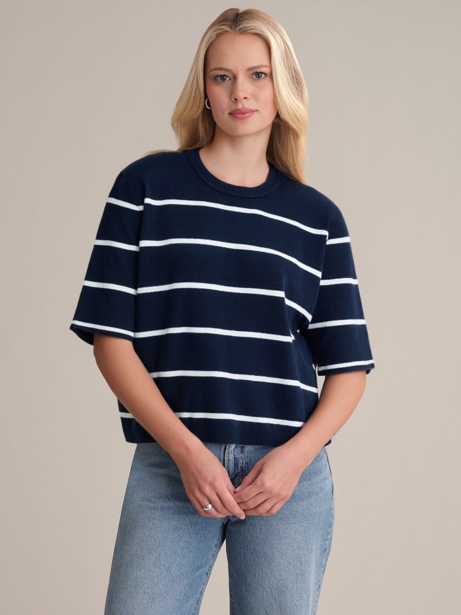 A woman with long blonde hair wears a 525 America classic crewneck navy blue and white striped short-sleeve shirt with light blue jeans, standing against a plain beige background.