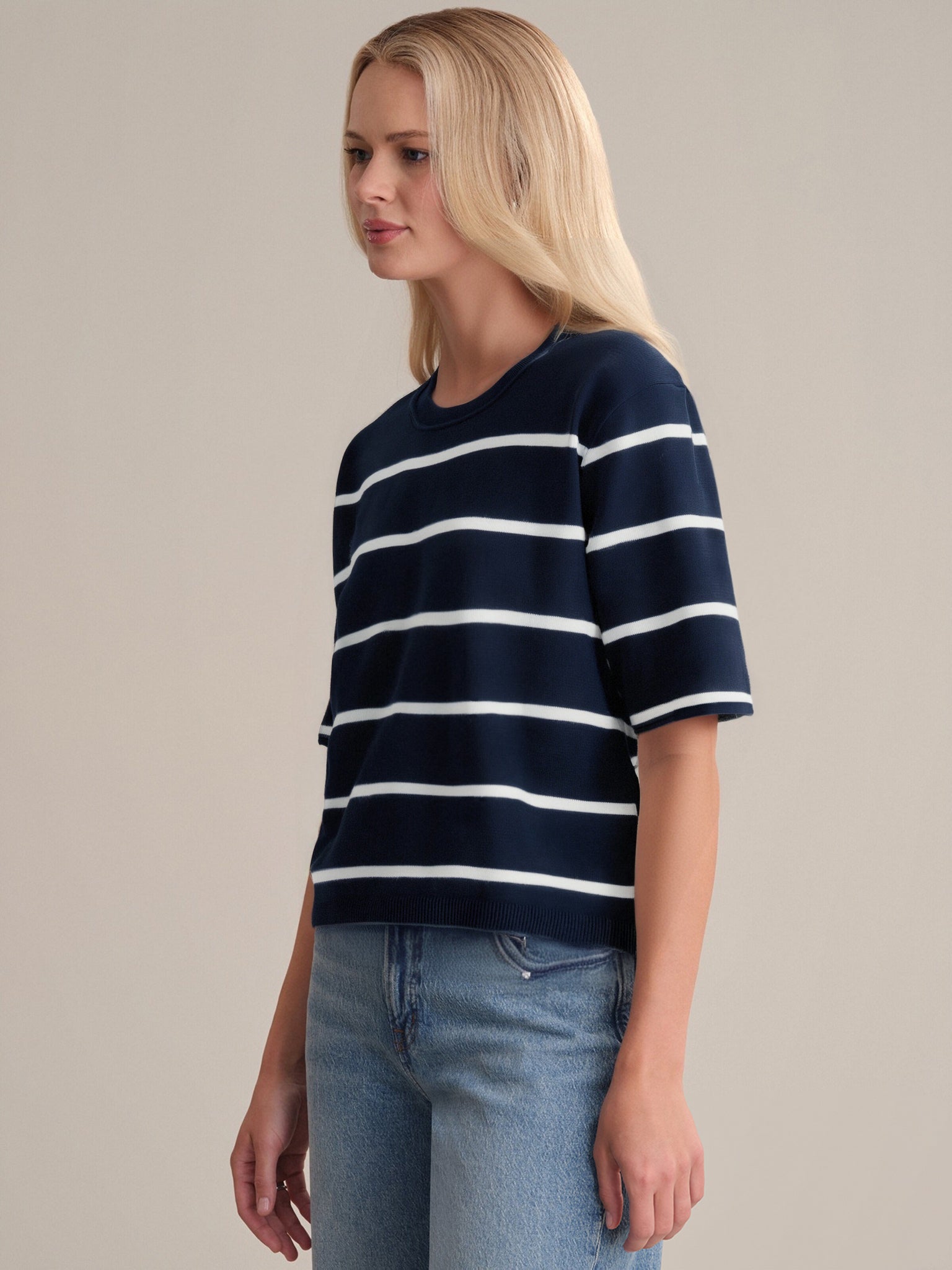 A person with long blonde hair models a 525 America navy blue short-sleeve crewneck shirt with white horizontal stripes and a boxy fit, paired with light blue jeans, standing before a plain, light-colored backdrop.