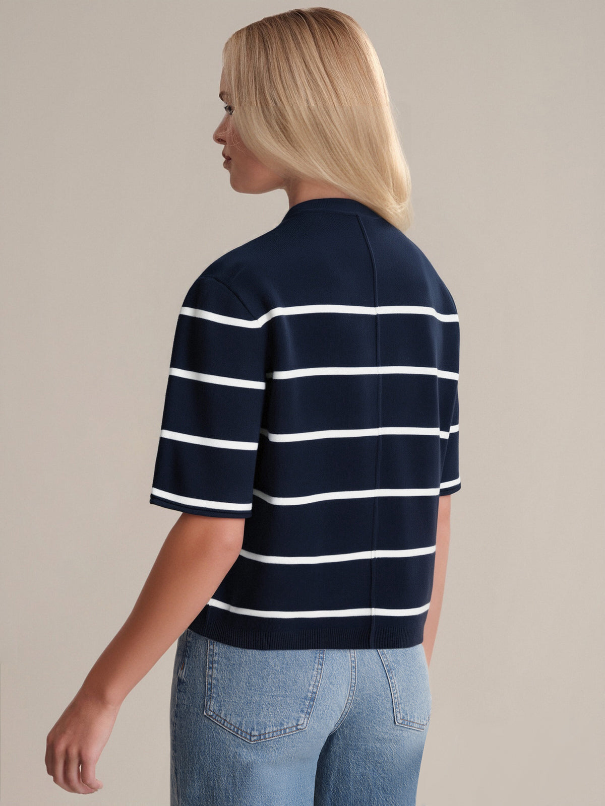 A person with straight, shoulder-length blonde hair is seen from behind wearing a 525 America navy premium cotton short-sleeve shirt with white horizontal stripes and light blue jeans, standing against a plain background.