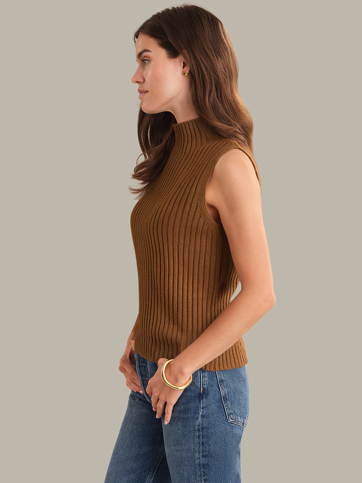 Woman wearing a brown ribbed sleeveless sweater and blue jeans against a neutral background