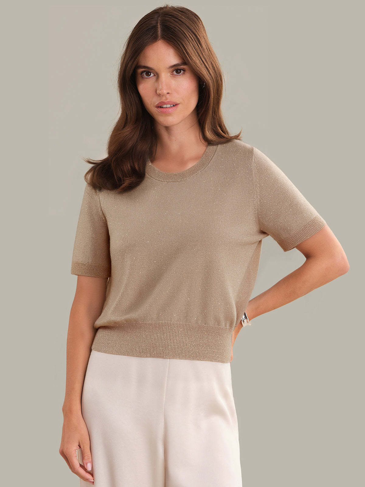 Woman wearing a beige sweater and white pants against a neutral background