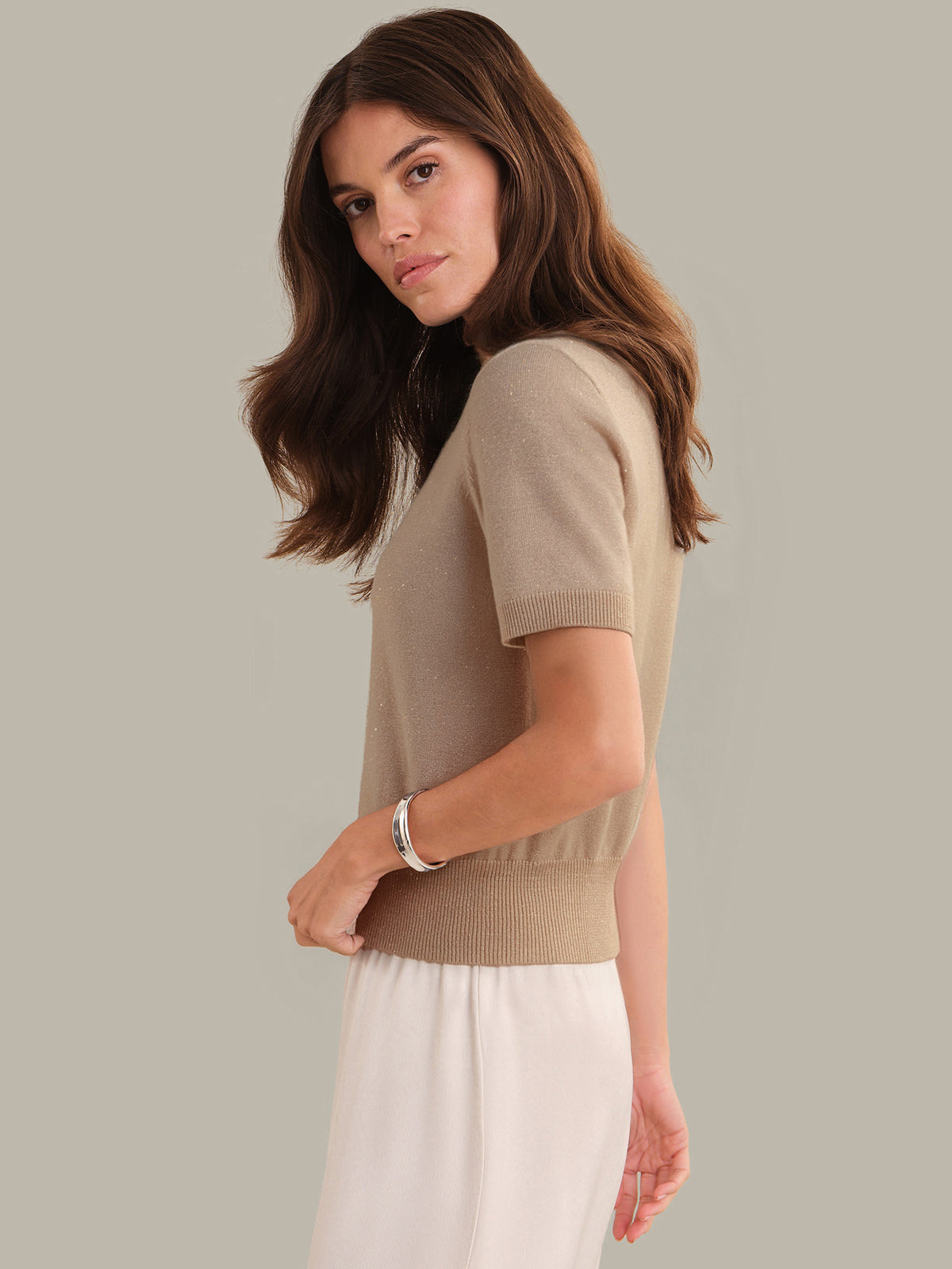 Woman wearing a beige sweater and white pants against a neutral background