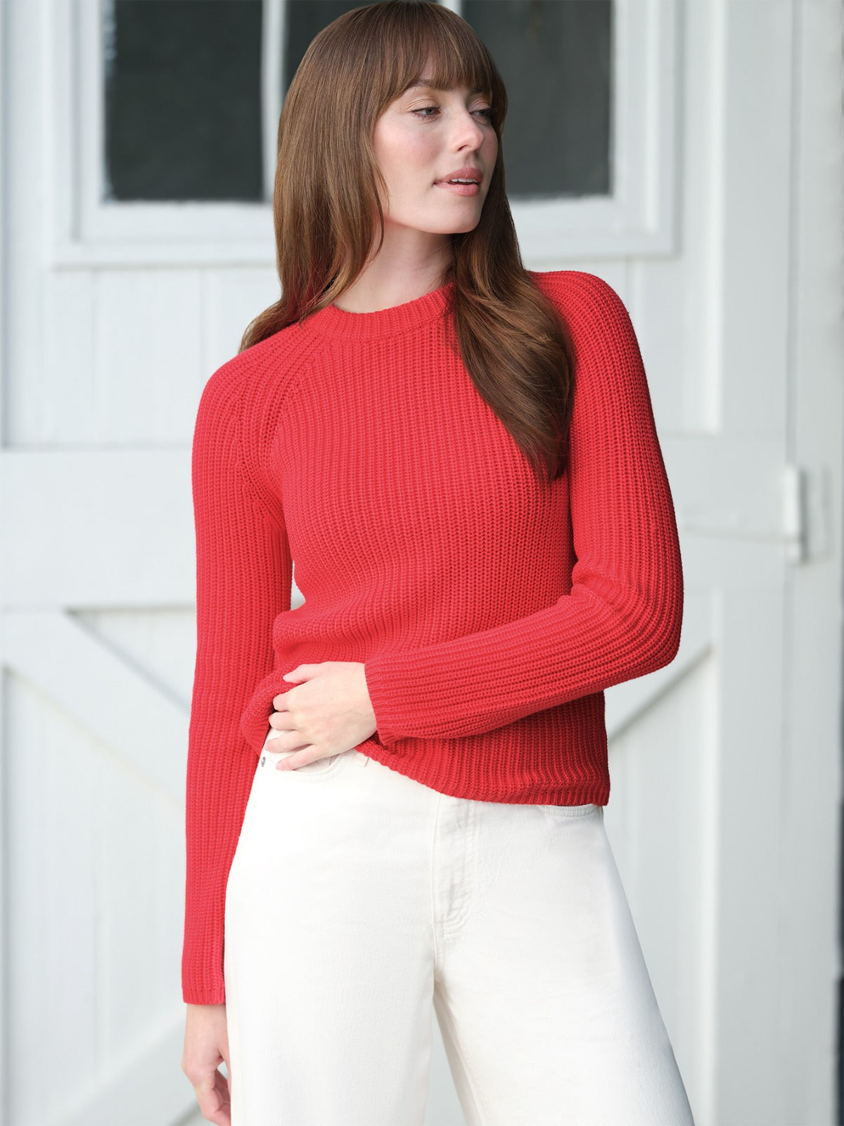 A person with straight brown hair wears a fitted red cotton shaker stitch sweater by 525 America and white pants, standing in front of a white wooden door and looking slightly to the side.