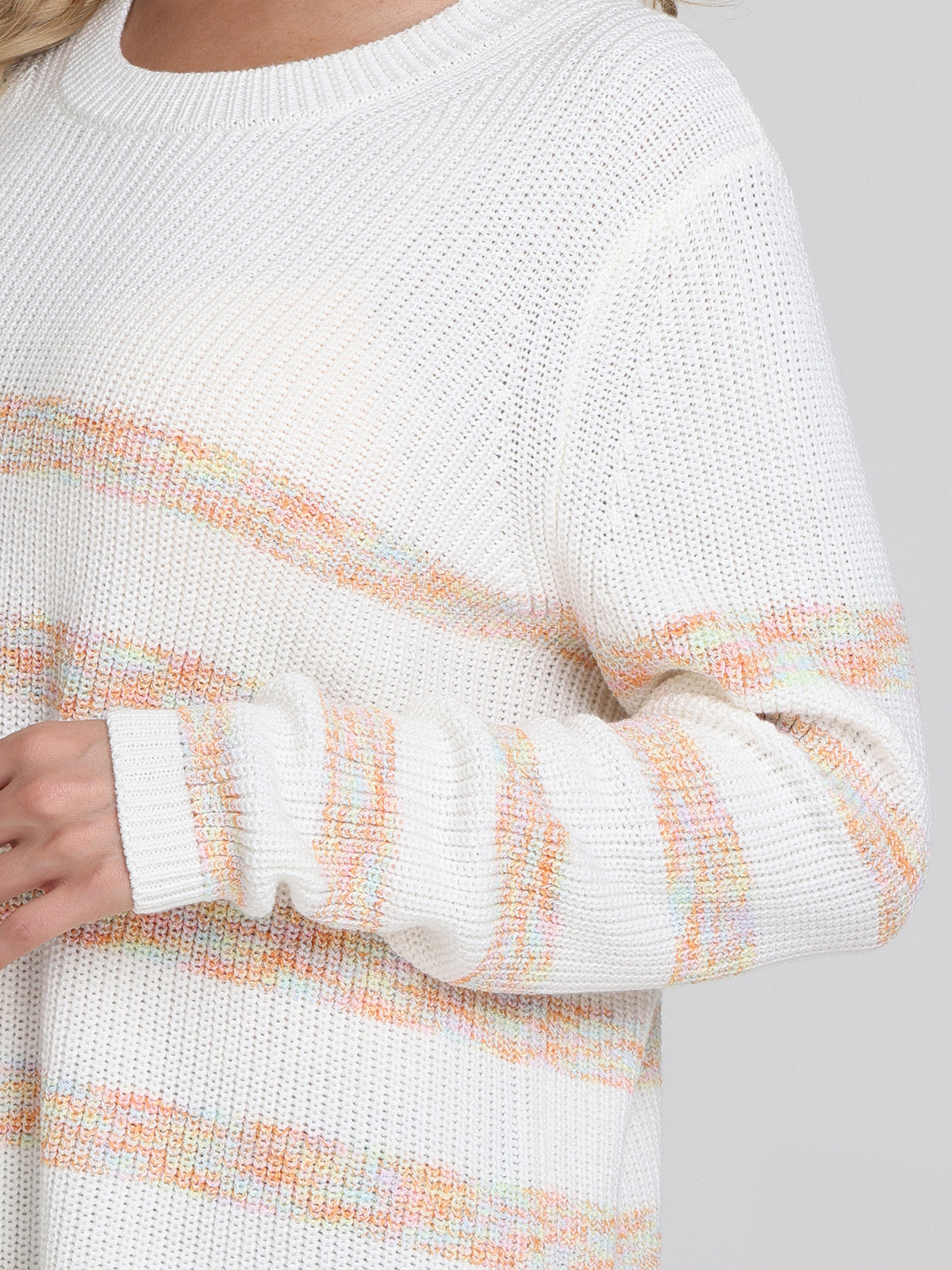 A person models a relaxed fit white knit sweater by 525 America, featuring pastel orange, yellow, and green stripes. Only the lower face, upper torso, and one hand appear against a plain light background.