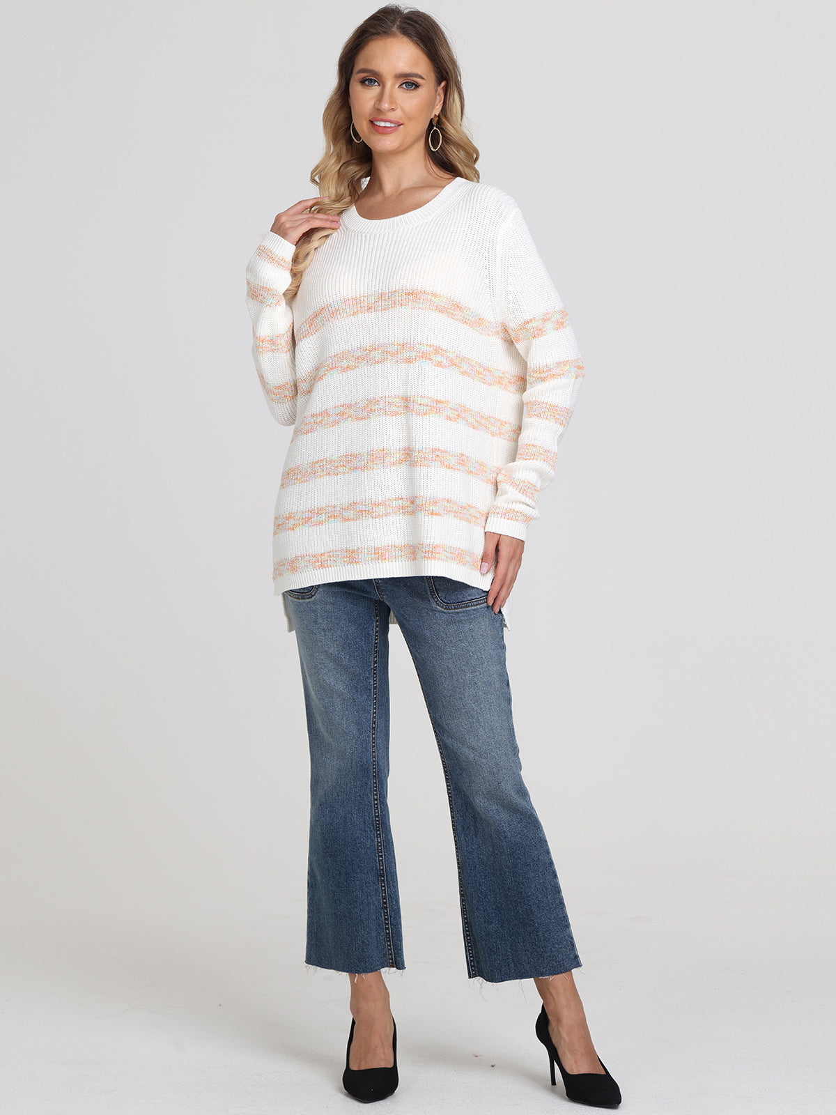 A woman wears a 525 America relaxed-fit white cotton shaker stitch sweater with pastel horizontal stripes, paired with blue cropped jeans and black heels. She has wavy hair, earrings, and smiles slightly against a plain light background.