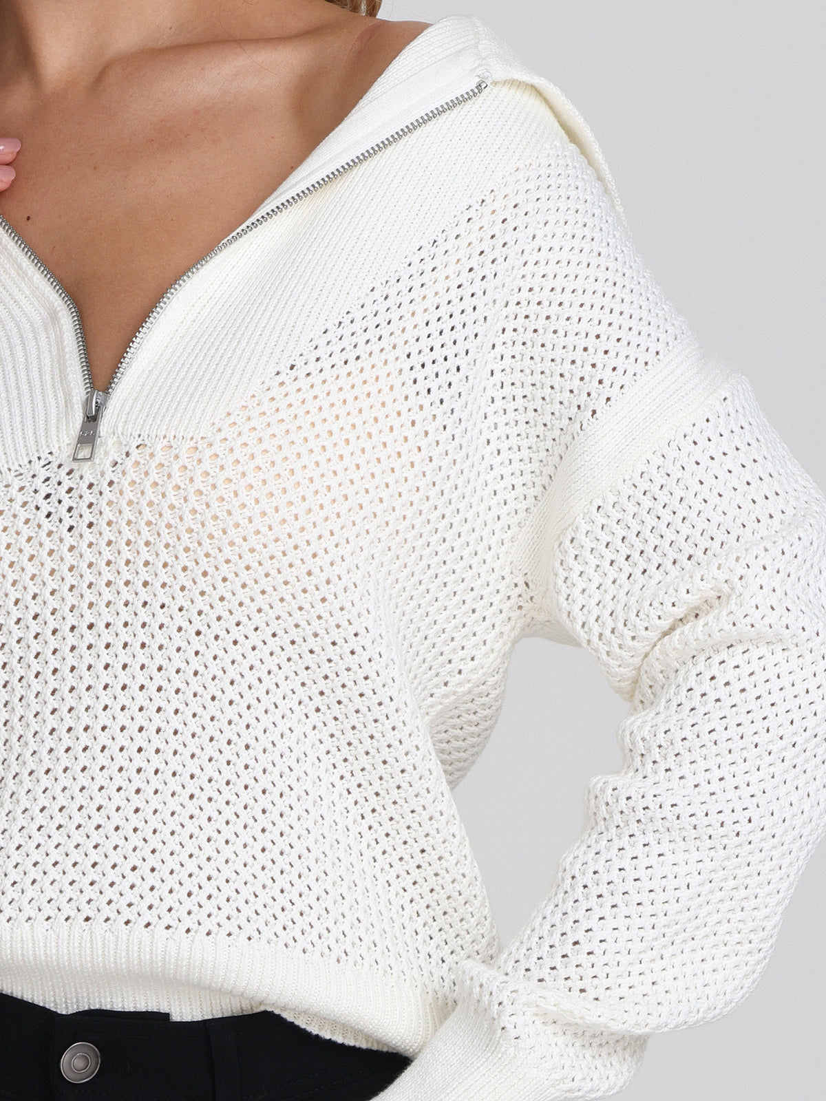 A person wears black pants and a relaxed-fit 525 America white long-sleeve pullover sweater with a perforated knit pattern and half-zip collar partially open, left hand near the neckline.