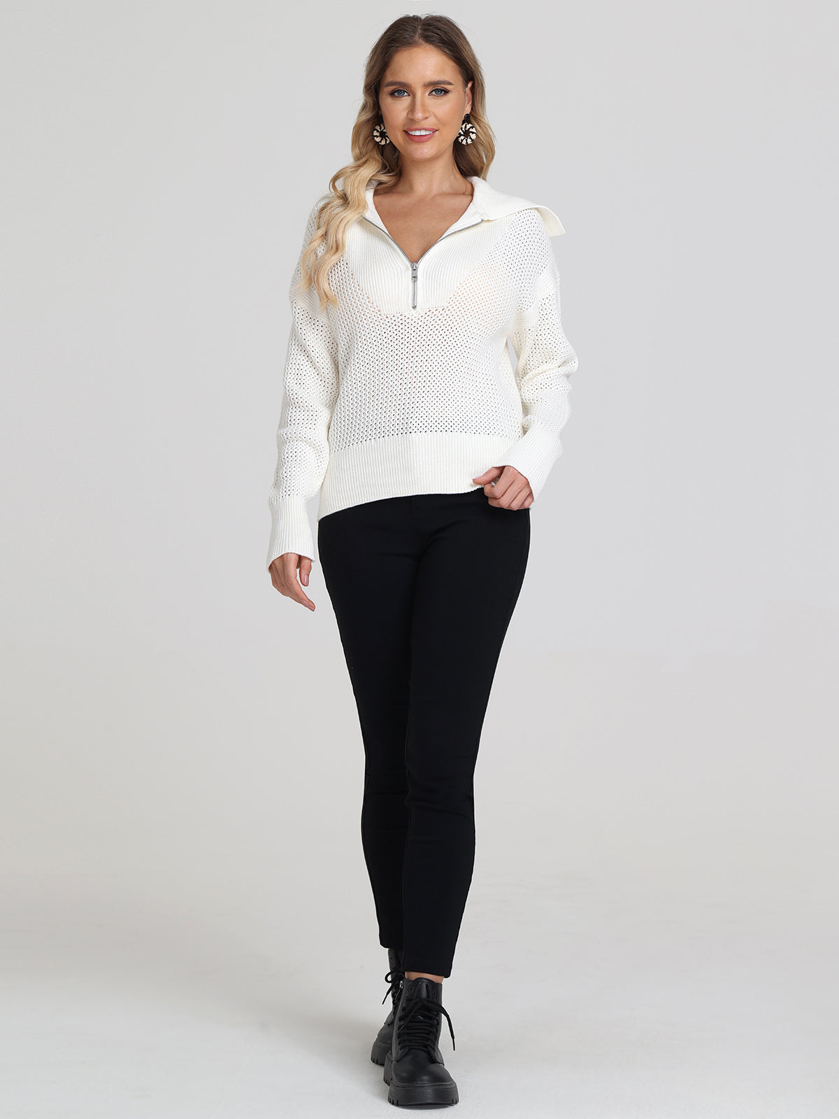 A woman stands against a plain background wearing a 525 America white knit pullover with a relaxed fit and half zipper, black pants, and black lace-up boots. She has long wavy hair, is smiling, and has one hand in her sweater pocket.