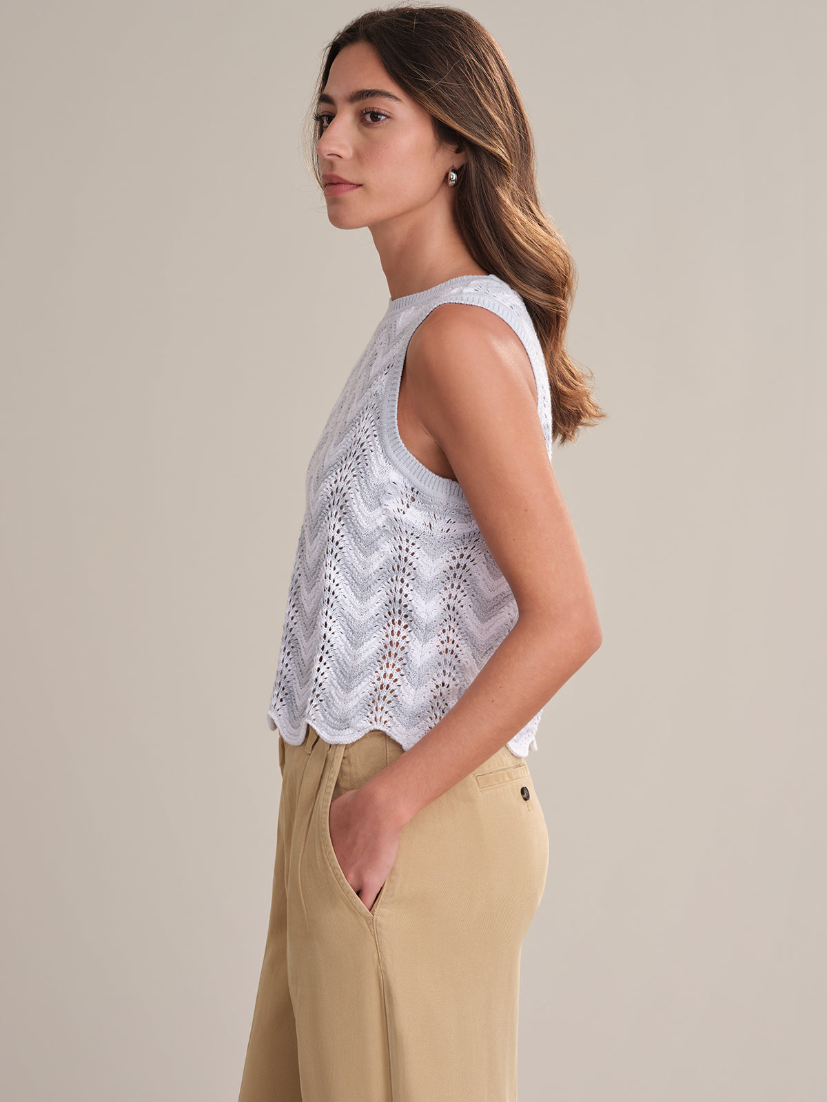 A woman stands in profile against a plain background, wearing a 525 America sleeveless white knit top featuring a chevron pointelle design and light beige high-waisted trousers. Her hands are in her pockets and her wavy hair is loose.