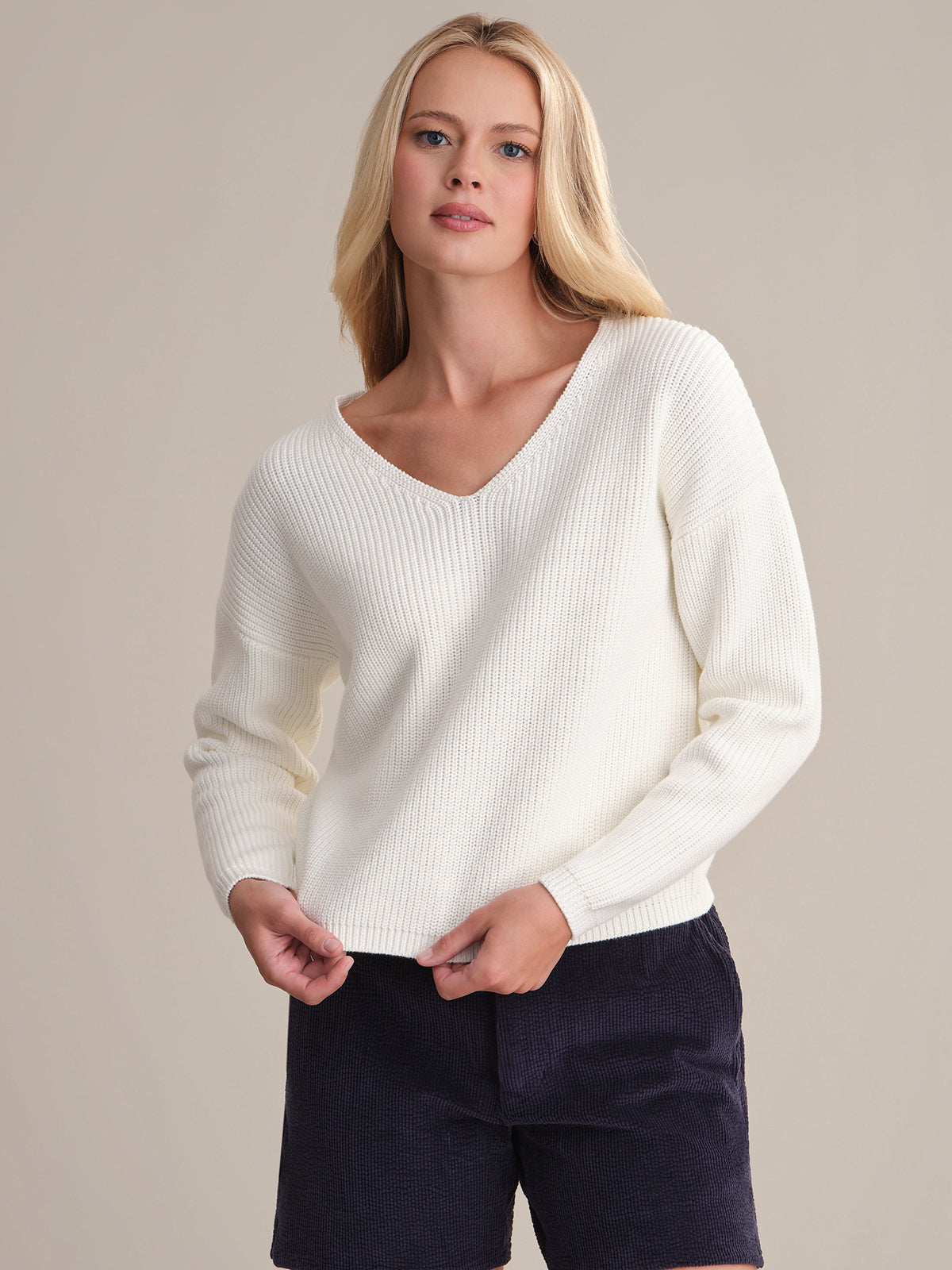 A person with long blonde hair wears the 525 America Ava V-Neck Sweater, crafted from 100% cotton with a shaker stitch design, paired with dark shorts. They are standing against a neutral background, looking forward in a relaxed posture.